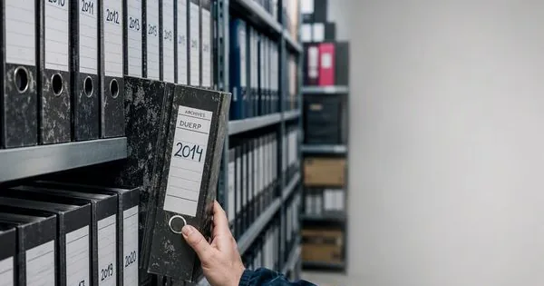 Main retirant d'une étagère d'archives un classeur étiqueté « Archives DUERP 2014 », au milieu de classeurs rangés par année